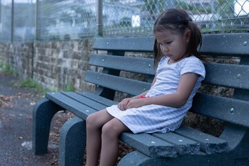 A sad little girl sitting alone outside on a bench depressed.