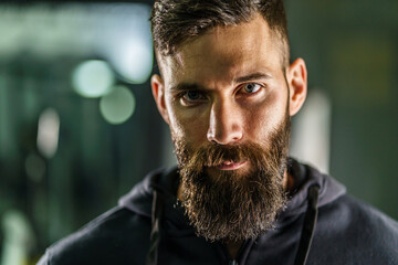 Front view close up portrait of young adult caucasian man with dark hair and beard in black shirt hoodie looking to the camera in dark - Confident modern male dramatic portrait with copy space