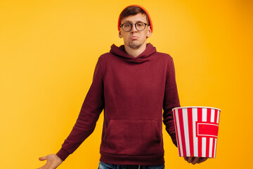 surprised young man wearing glasses and red hat watching movie holding bucket of popcorn on yellow...