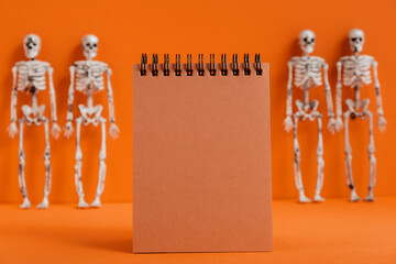 Empty black notebook on an orange background with skeletons in blur. Modern Halloween or Day of the Dead concept. A minimalistic holiday design for Halloween preparations. Copy space.