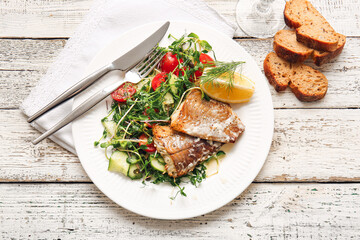 Plate with tasty baked cod fillet and vegetables on light wooden background