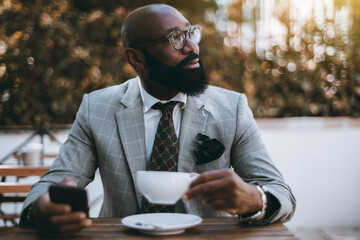 A portrait of a mature black senior entrepreneur sitting in a street cafe with a cup of delicious tea in one hand and a smartphone in another; African American businessman having a coffee break - Powered by Adobe