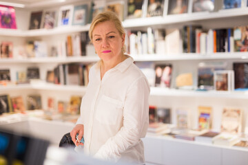 Portrait of handsome adult mature lady choosing new books on shelves in museum shop