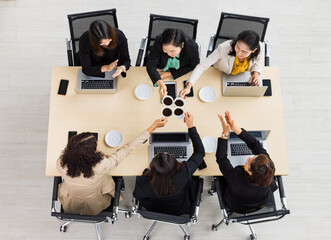 Top view of six businesswomen sitting together around wooden conference table , cheering and clinking coffee mug together with laptops and tablets on table in office. Concept for business meeting