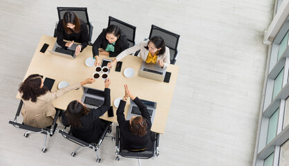 Top view of six businesswomen sitting together around wooden conference table , cheering and clinking coffee mug together with laptops and tablets on table in office. Concept for business meeting