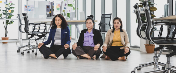 Three asian business women sitting on the floor in office with multiple glass windows meditating beside conference tables