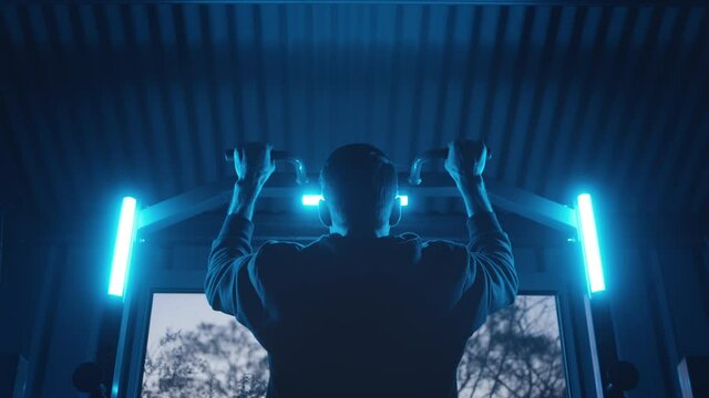 A young man in black doing chin ups in a gym room with blue lights around him. A person working out doing exercises in a dark and moody setting.
