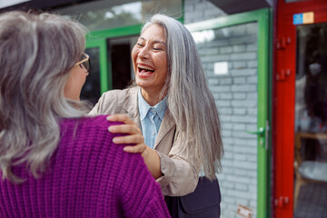Emotional mature Asian lady laughs meeting grey haired best friend on city street on autumn day. Long-time friendship relationship