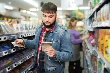 Positive bearded guy choosing chocolate on shelves in grocery store