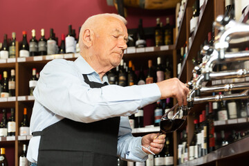 Elderly seller pouring wine from wine column in liquor store