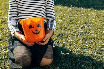 Halloween concept. A girl in a suit sits on the grass and holds an orange pumpkin bucket in her...