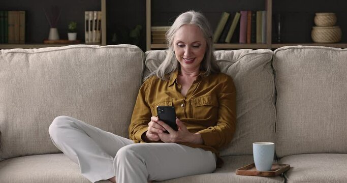 Attractive mature grey-haired woman relaxing on sofa with smartphone, read media news, learns new cool mobile application, spend free leisure at home using modern wireless tech and internet concept