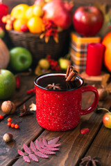 red mug with mulled wine. Cinnamon sticks stick out of the cup and a star of star anise floats. Fruits and spices are all around on a wooden table