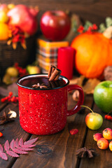 red mug with mulled wine. Cinnamon sticks stick out of the cup and a star of star anise floats. Fruits and spices are all around on a wooden table
