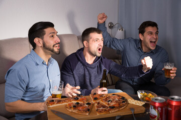 Exalted male friends watching tv together at home, enjoying beer and pizza