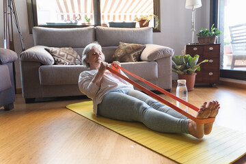 Older woman exercising with an elastic band lying on a mat on the floor of her living room at home