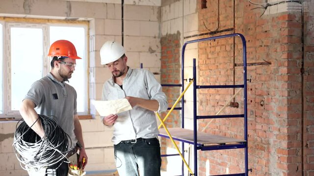 In the middle of the room being renovated, two construction workers in helmets are standing and discussing.