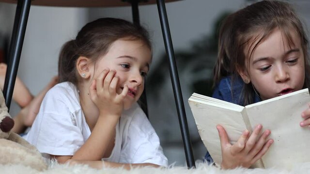 Little girls' sisters have fun reading a book lying on the floor at home.