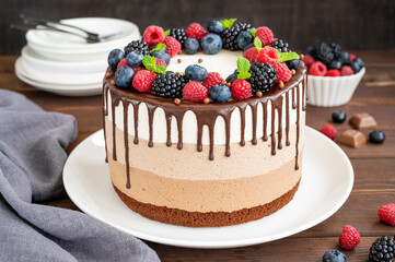 Three chocolate mousse cake with chocolate glaze, blueberries, raspberries and blackberries on top on a white plate on a dark wooden background. Cake with three layers of different taste. Copy space.