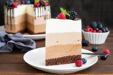 Three chocolate mousse cake with chocolate glaze, blueberries, raspberries and blackberries on top on a white plate on a dark wooden background. Cake with three layers of different taste. Copy space.