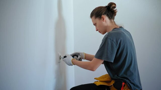 Professional repairwoman with tool belt fixing socket with a screwdriver on a white wall. Young woman doing home repairs.