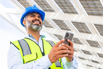 Ethnic smiling supervisor in hardhat with smartphone against solar power station