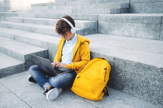 Child caucasian teen boy in yellow hoodie with headphones sitting outdoors on grey stairs using laptop. Remote or distant learning on the go. Blogging surfing. Homeschooling. Lockdown. Video call.