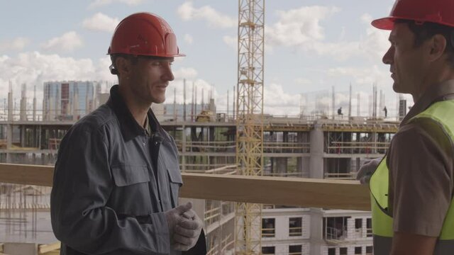 Medium of two male Caucasian construction workers wearing helmets standing in foreground of multistory building under construction, talking on sunny day