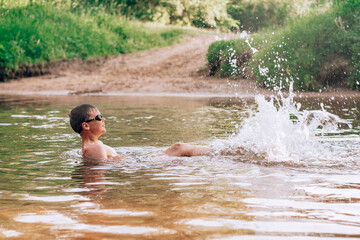 Cheerful funny boy 7-10 lies in river wearing goggles for swimming, splashes in rays of sun