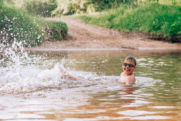 Cheerful funny boy 7-10 lies in river wearing goggles for swimming, splashes in rays of sun
