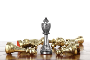 Silver king among fallen golden chess pieces on wooden board against white background