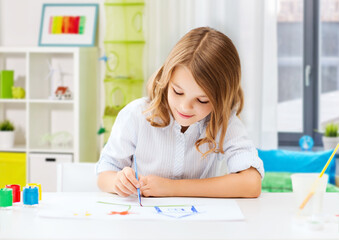 hobby, art and childhood concept - happy smiling girl with brush and colors drawing picture over home room background