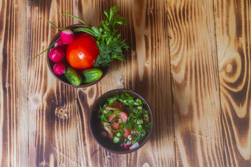 Vegetable salad in a clay plate of tomatoes, cucumbers, radishes, dill, onions, parsley on a wooden background