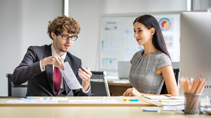 Handsome caucasian man as executive manager talk and teach beautiful corporation colleague to analyze interesting paper of business portfolio at working desk in meeting room of company office