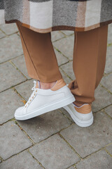 Closeup photo of beautiful fashionable woman wearing checkered long coat, beige pants, white blouse and sneakers.