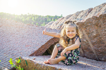 joyful girl sitting on the stones shows her thumb up, the sun illuminates her.