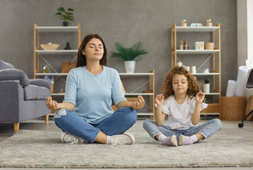 Calm young Caucasian mom and 7s daughter sit on floor with mudra hands meditate relieve negative...