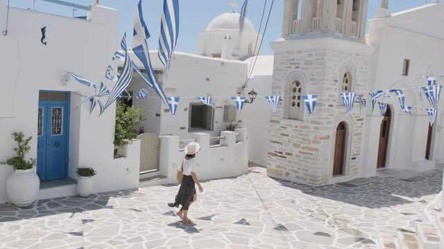 Woman with hat walking in slow motion in the main square of a small town of white houses and Greek flags in the island of Paros.