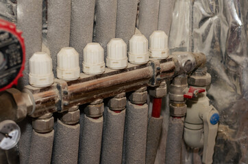 Floor heating manifold installed on the wall. Insulated water supply and circulation pipes. Red electric water pump. Thermometer. Inside the room.