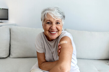 Portrait of a senior woman proudly showing her arm with bandage after getting vaccine. Mature white haired woman sitting against bright background after receiving coronavirus vaccination, thumbs up. - obrazy, fototapety, plakaty