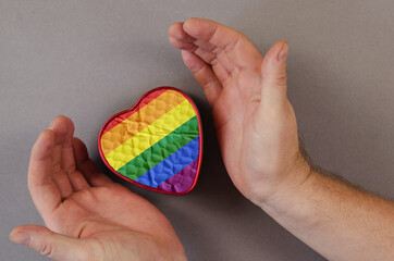 An adult male embraces a red small metal heart-shaped box. Hands hold a closed gift with a rainbow flag. Top view. Selective focus.