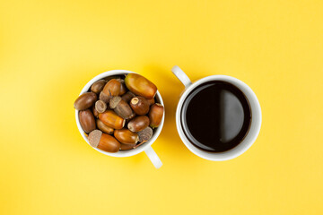 Two white cups with alternative coffee without caffeine and ripe acorn on yellow background
