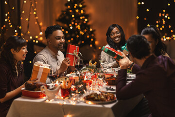 holidays, party and celebration concept - multiethnic group of happy friends having christmas dinner and giving presents at home