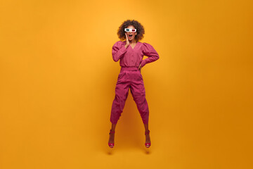 photo of straight happily surprised African jumping in place with legs apart on yellow background. girl in 3D glasses with flying hair is dressed in bright festive outfit and open sandals with heels.