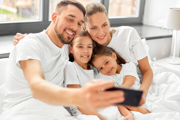 family and people concept - happy mother, father and two daughters taking selfie with smartphone in bed at home