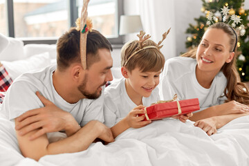 family, winter holidays and people concept - happy mother, father and little son in pajamas with christmas gift in bed in morning