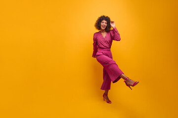 radiant swarthy woman in purple suit and pink shoes on isolated background. curly brown-haired...