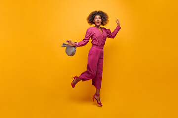 Fototapeta na wymiar irresistible sophisticated lady jumping elegantly posing on isolated yellow background. her lush curly hair flies to sides. dressed in purple suit with handbag in her hands.