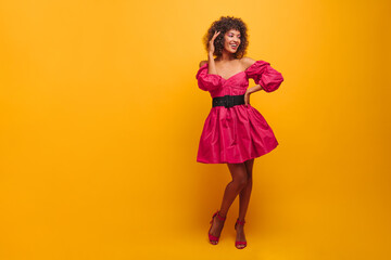 full-length glamorous lady elegantly posing for photo looking to side against background of mustard color. young woman in crimson dress with wide black belts and open shoes with heels.