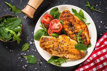 Grilled chicken fillet at white plate with green leaves and vegetables. Top view at black stone table.